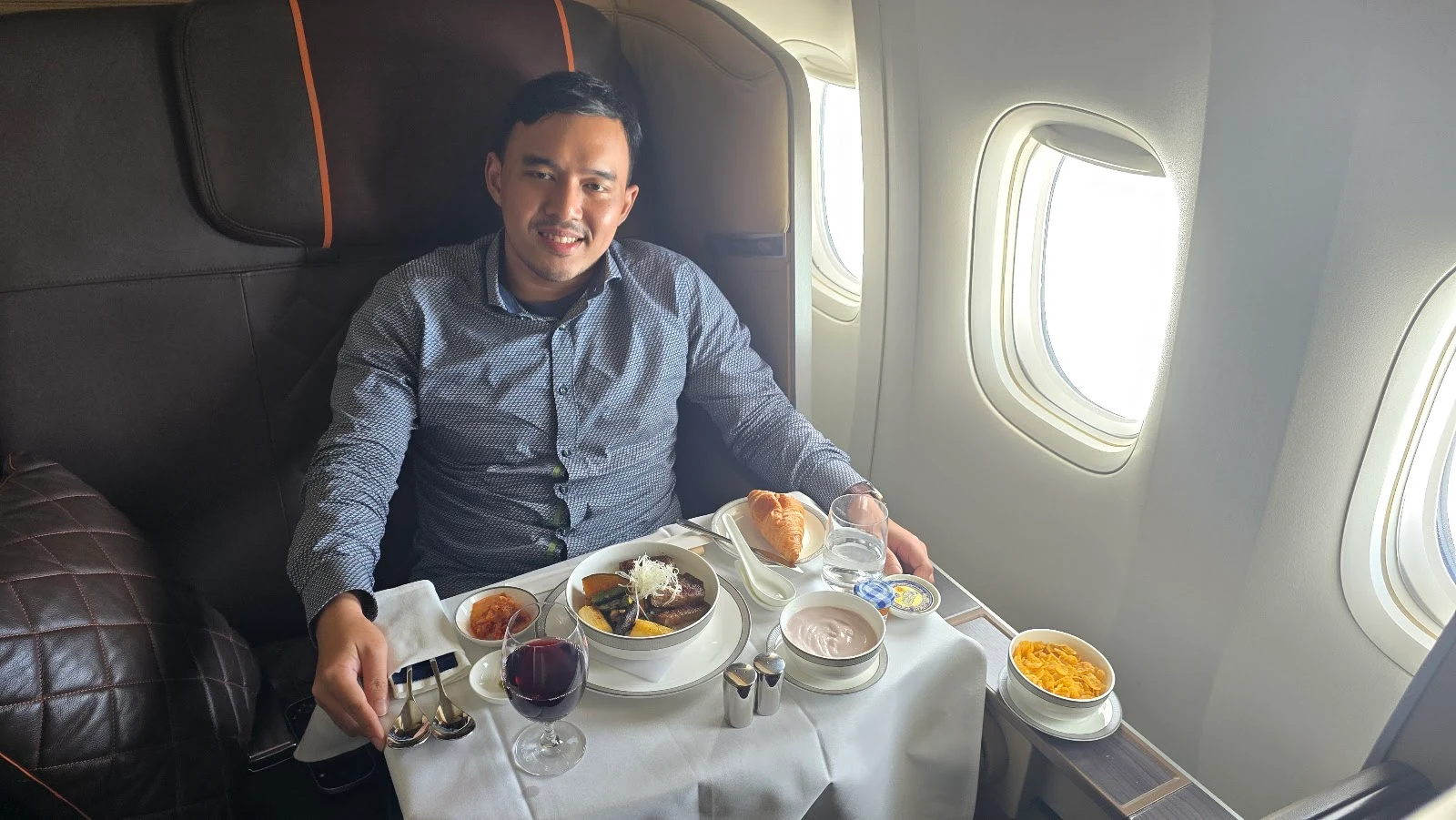 Adjie smiling at his seat in Singapore Airlines First Class, with beef stew, a croissant and a glass of red wine on the table